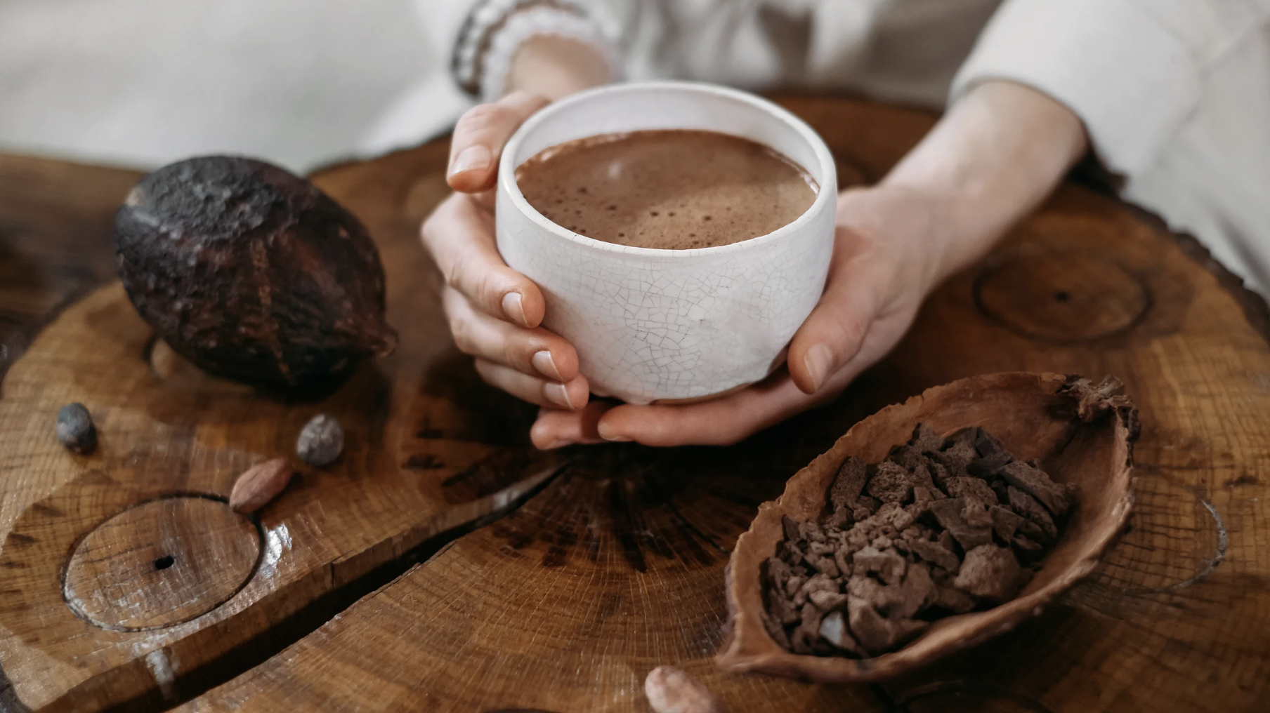 Foto einer Hand, die eine Tasse gefüllt mit Zeremoniekakao hält, dazu Kakobohnen auf einer schönen Holzunterlage